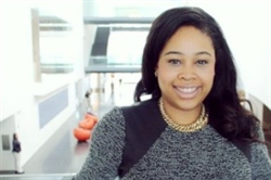 [ai] A woman with long dark hair and a smiling expression wears a black and gray sweater with a gold necklace, posing indoors in a modern building with bright lighting.